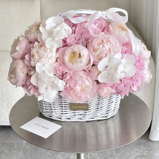 Pink preserved peonies and hydrangeas with white orchids in a handled basket on a table