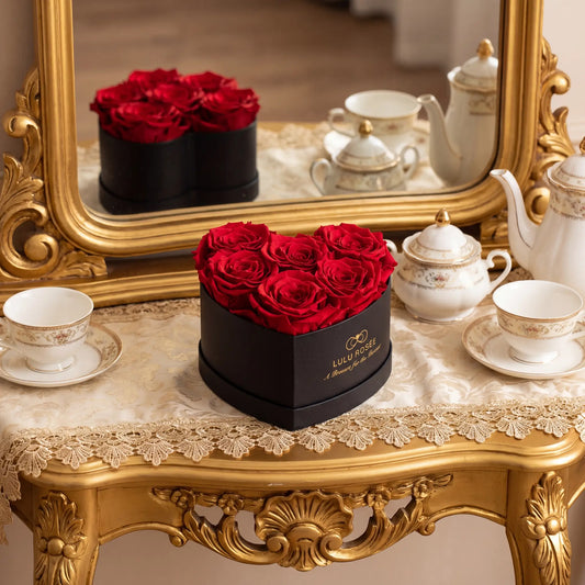 LULU ROSÉE heart shaped black gift box with preserved deep red roses, placed on vintage golden carved table with lace tablecloth, antique porcelain tea set and ornate gold mirror, luxury romantic preserved rose arrangement