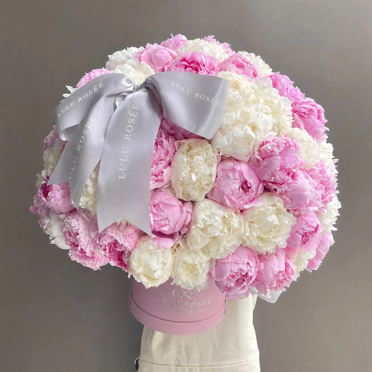 A round, lush bouquet of pink and white peonies in a pink round box, tied with a wide light gray satin ribbon printed with the brand name LULU ROSÉE