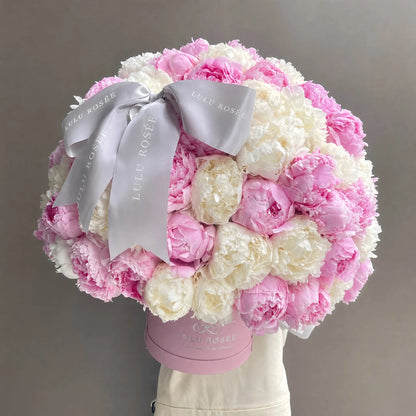 A round, lush bouquet of pink and white peonies in a pink round box, tied with a wide light gray satin ribbon printed with the brand name LULU ROSÉE