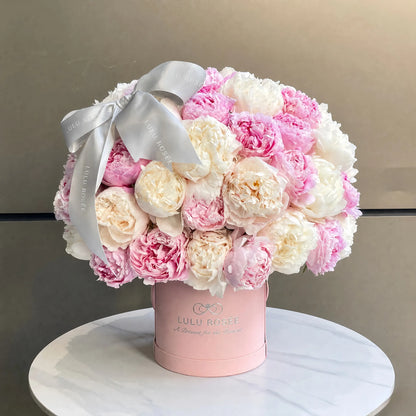 A round bouquet of pink and cream peonies in a light pink cylindrical box with a silver bow, displayed on a white marble round table against a neutral gray background. The box is branded LULU ROSÉE