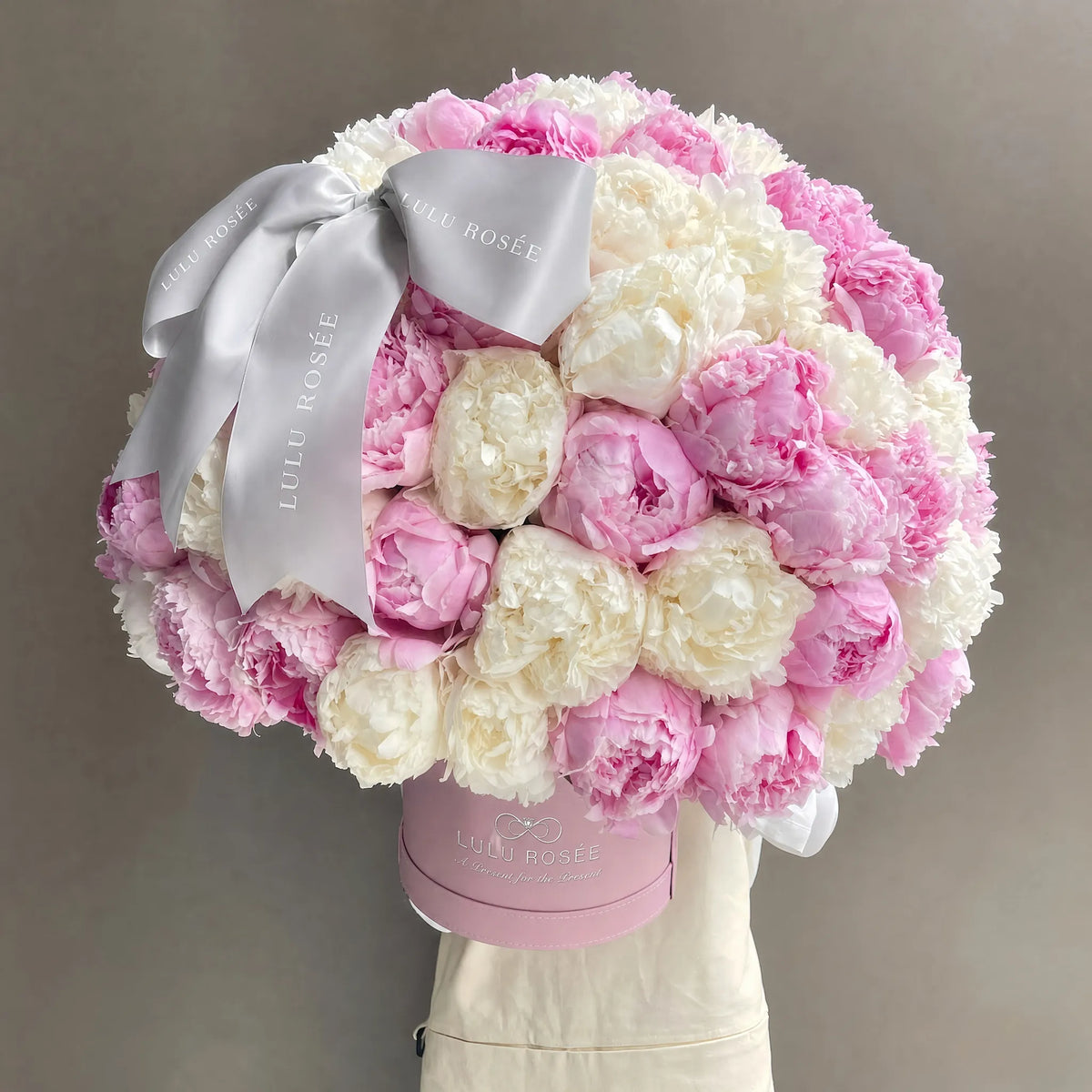 A large round bouquet of pink and white preserved peonies in a pink round box, tied with a wide silver satin ribbon printed with the brand name 'LULU ROSÉE'