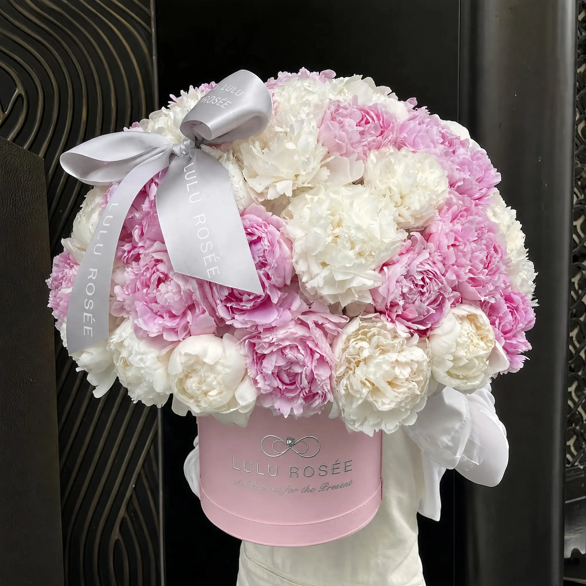 A round floral arrangement of pink and white peonies in a pink Lulu Rosée box, tied with a white satin bow and ribbon, set against a dark patterned background