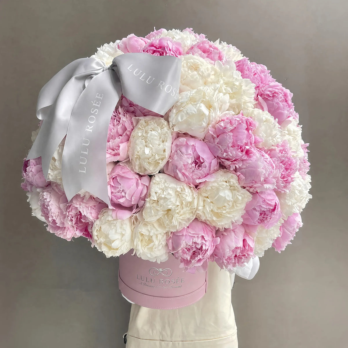 A round bouquet of pink and white peonies arranged in a light pink round box, tied with a wide white satin ribbon printed with the brand name 'LULU ROSÉE'