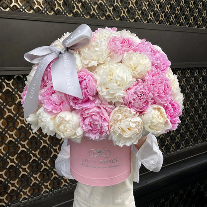 A large bouquet of pink and white peonies arranged in a round pink box with a white ribbon bow. The box is branded LULU ROSÉE and is held by a person wearing a white shirt, set against a dark patterned wall