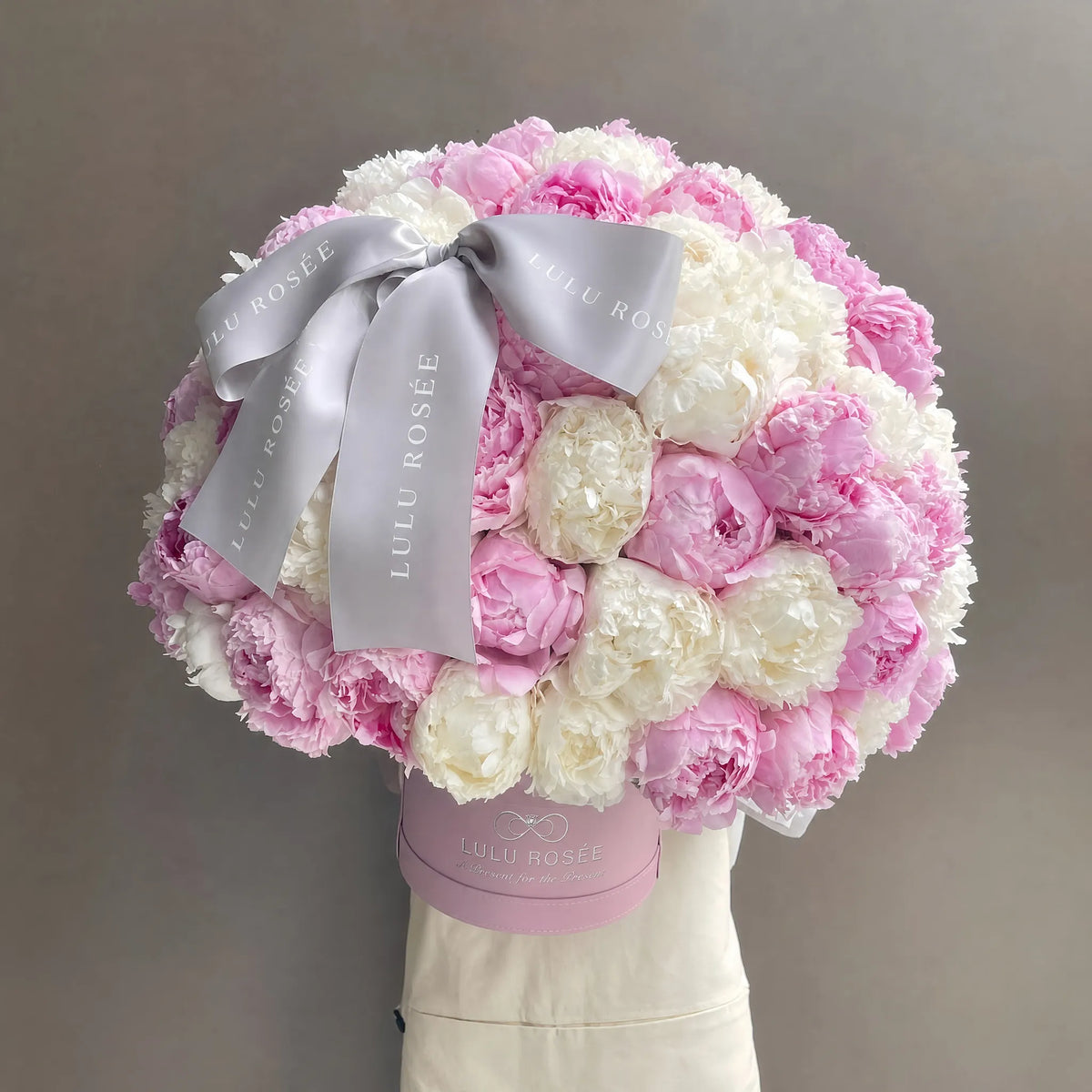 A round floral bouquet of pink and white peonies in a pink box, tied with a wide silver satin ribbon printed with 'LULU ROSÉE', displayed on a mannequin torso against a plain taupe background