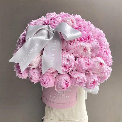 A large bouquet of light pink peonies arranged in a round pink box, tied with a wide silver satin bow. The box is branded 'LULLI ROSÉE' and is displayed against a plain gray background.