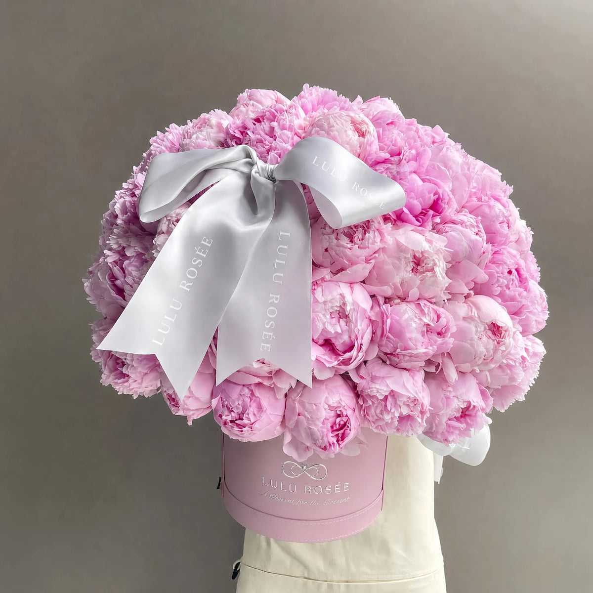 A large bouquet of light pink peonies arranged in a round pink box with a wide white satin bow. The box is branded 'Lulu Rosée' and is displayed on a cream-colored mannequin torso against a plain gray background
