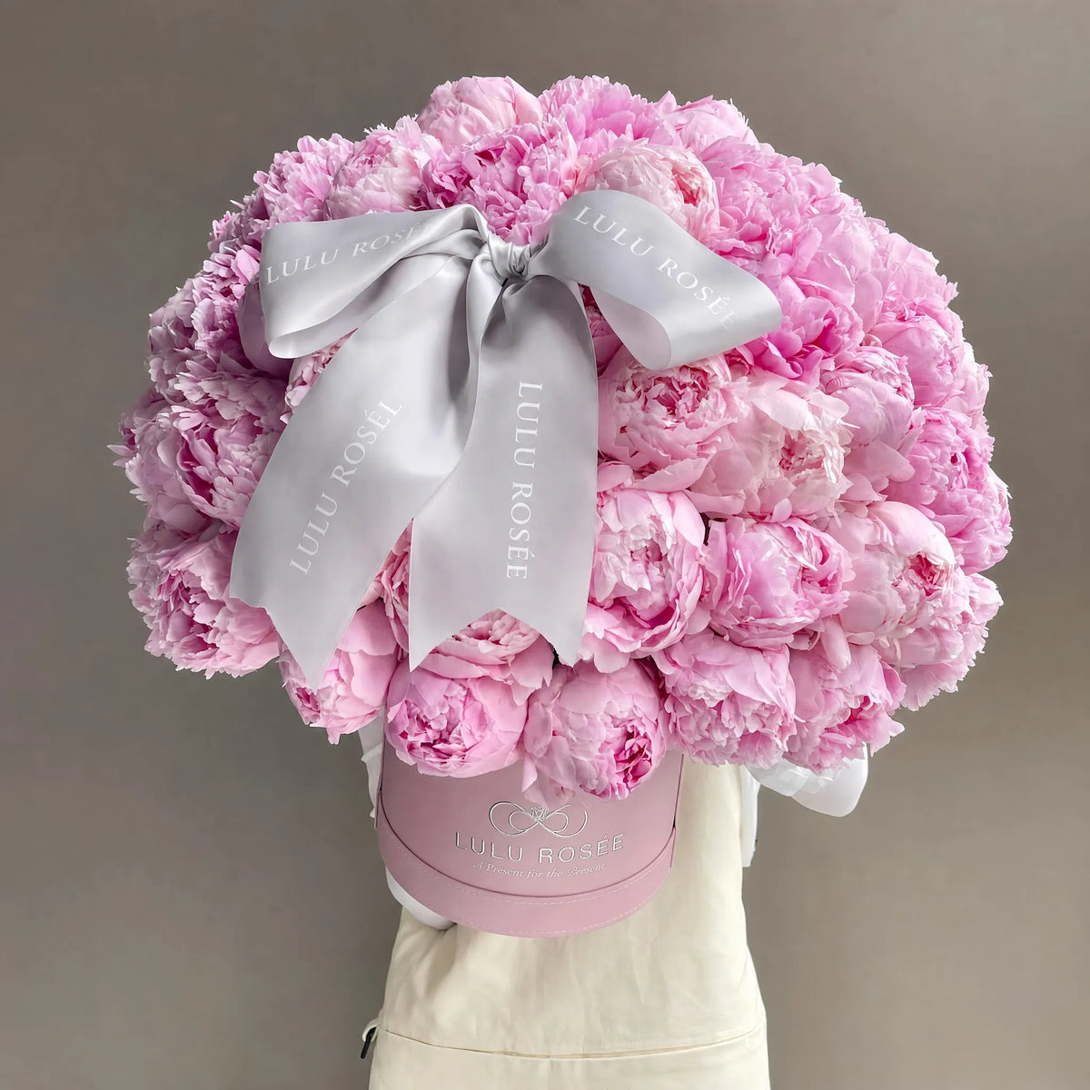 A large bouquet of pink peonies arranged in a round pink box, tied with a wide white satin ribbon printed with the brand name 'LULU ROSÉE'