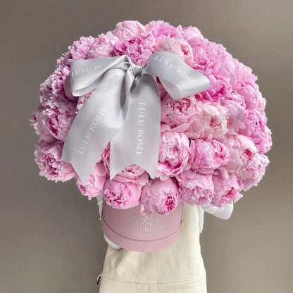 A large bouquet of pink peonies arranged in a round pink box, tied with a wide white satin ribbon printed with the brand name 'LULU ROSÉE'