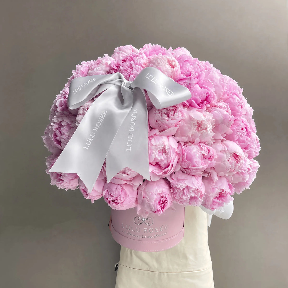 A large bouquet of light pink peonies arranged in a round shape, tied with a wide white satin bow printed with the brand name 'LULU ROSÉE', presented in a pink round box against a neutral gray background