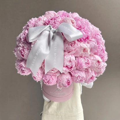 A large round bouquet of light pink peonies arranged in a round pink box, tied with a wide white satin bow printed with the brand name LULU ROSÉE