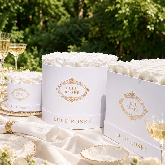 White rose boxes with gold branding arranged with crystal glasses and table linens in a garden setting.