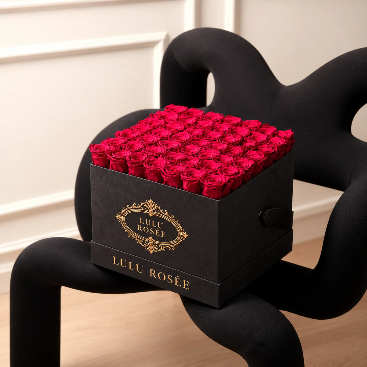 Red preserved roses in a Grey Suede LULU ROSÉE large square box beside a sculptural chair.