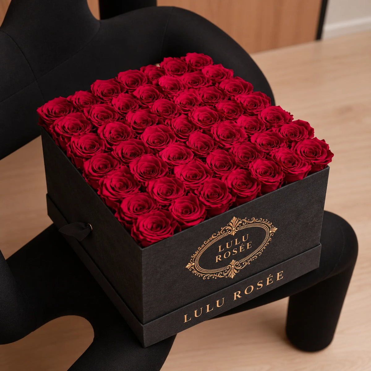 Red preserved roses in a Grey Suede LULU ROSÉE large square box styled on a sculptural black chair.