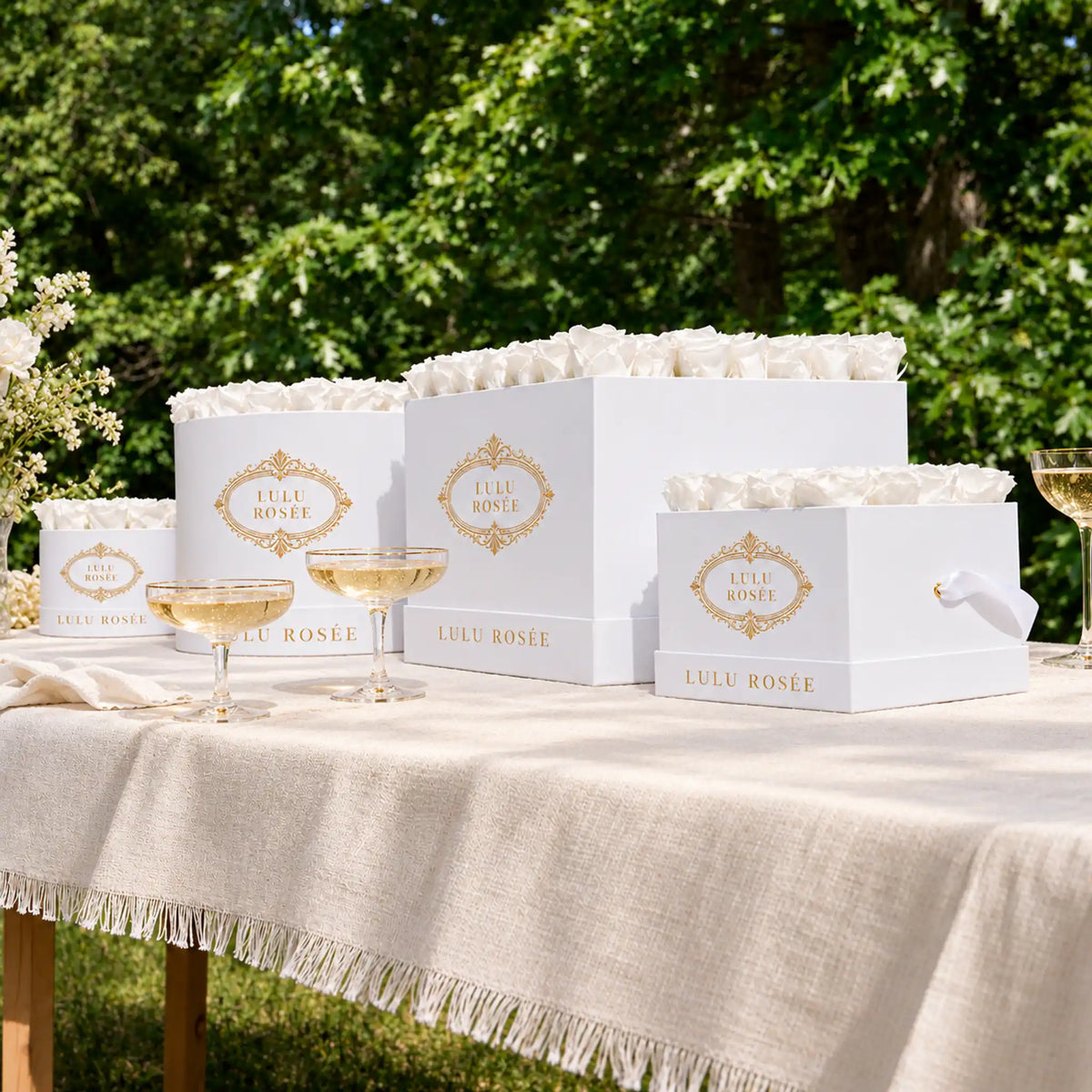 White preserved rose boxes by LULU ROSÉE displayed on a garden table with champagne.
