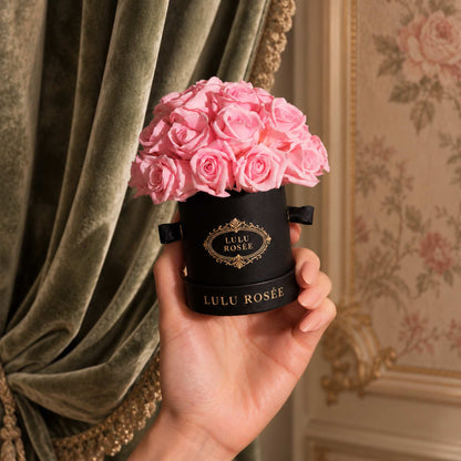 A hand holds a black classic Mini Le Plein box filled with light pink eternal roses.
