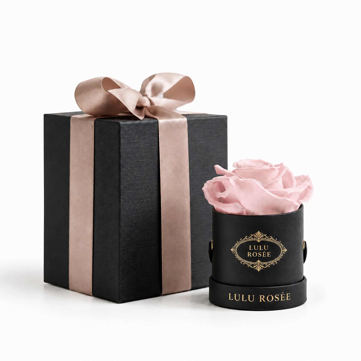 A light pink eternal rose in a black classic Mini Round box beside a wrapped gift box.