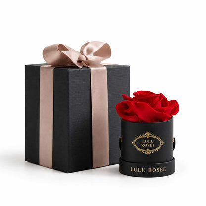 A red eternal rose in a black classic Mini Round box beside a wrapped gift box.