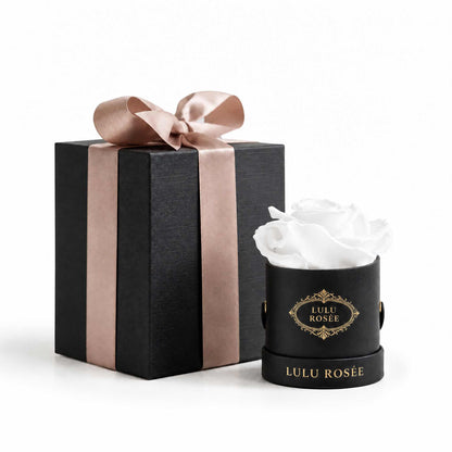 A white eternal rose in a black classic Mini Round box beside a wrapped gift box.
