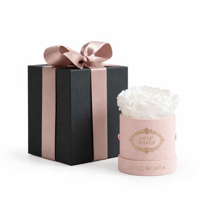 A white eternal rose in a blush suede Mini Round box beside a wrapped gift box.