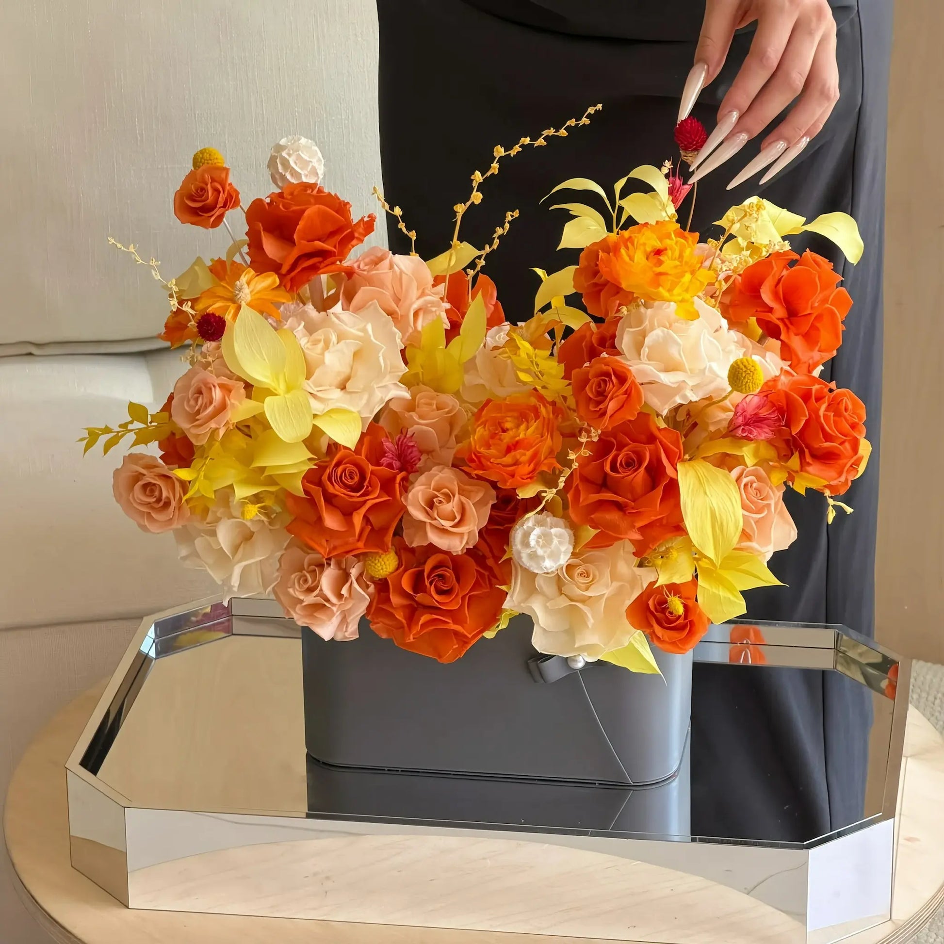 LULU ROSÉE Close-Up of Sunset Orange Parisian Bloom Garden in a vase on a reflective surface with a person in the background