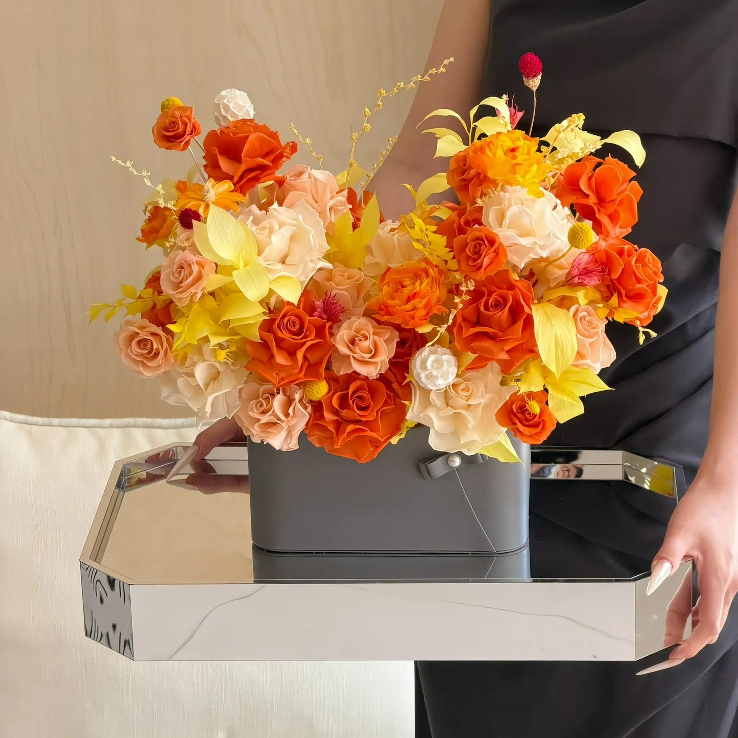 LULU ROSÉE Sunset Orange Parisian Bloom Garden Preserved Flower Arrangement in a gray vase on a table with a person partially visible