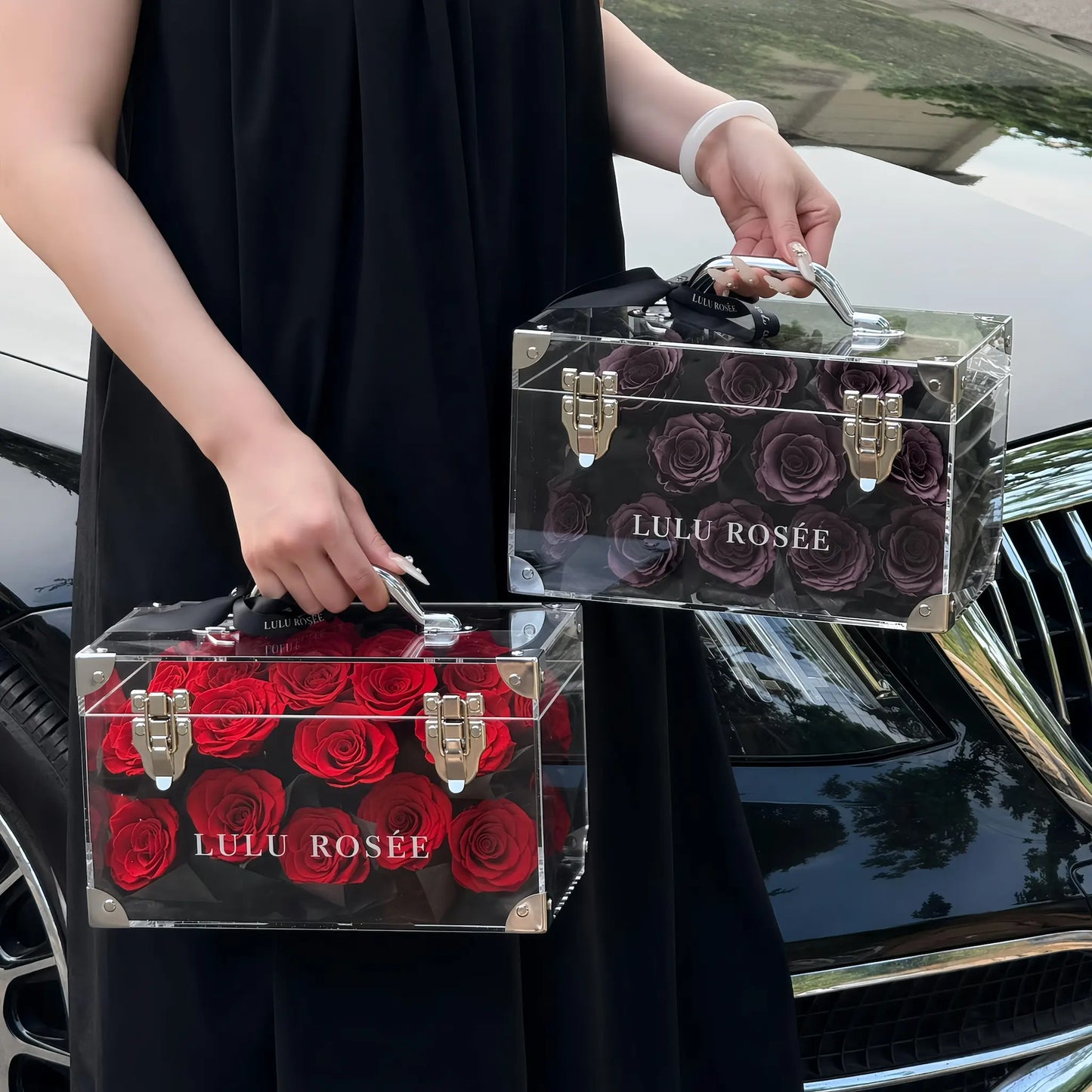 LULU ROSÉE Petite Eternal Acrylic Handbag Rose Box, small transparent acrylic handbag-shaped gift box, wine red and dark purple two box handheld lifestyle shot with car background, customizable flower color options, mini luxury everlasting floral souvenir for birthday, anniversary & romantic gift giving