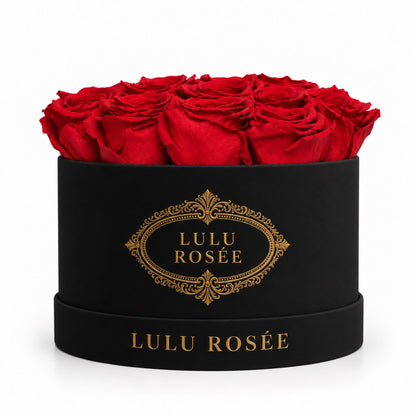 Red preserved roses in a black round box with ornate gold logo detailing.