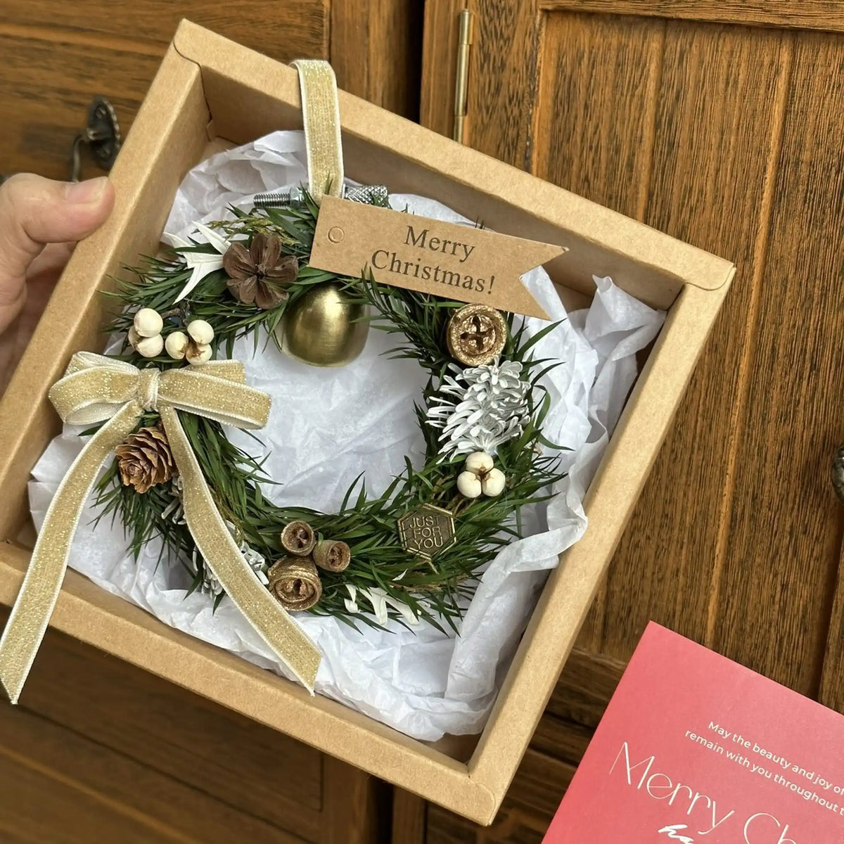 Small Christmas bell wreath nestled in tissue paper inside a gift box beside a card.