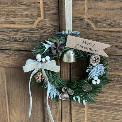 Green Christmas wreath with a bell, cream ribbon and festive tag hanging on a wooden door.