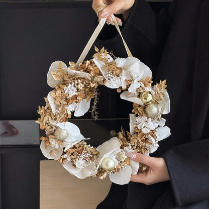 White and gold preserved flower Christmas wreath held against a dark background.