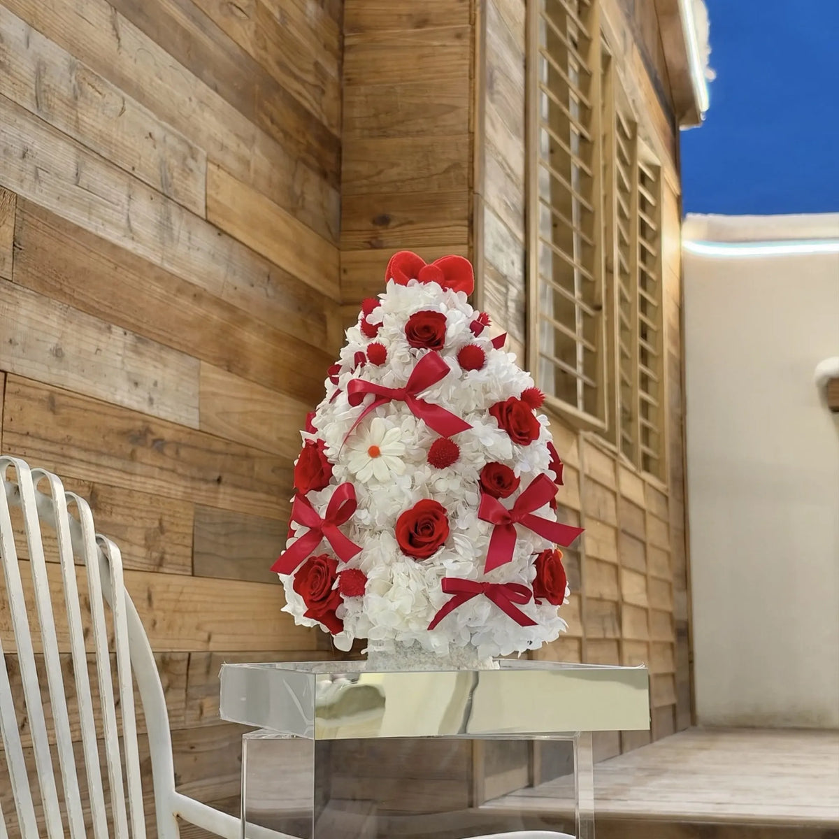 Red and white preserved flower Christmas tree on a mirrored base beside a wooden wall.