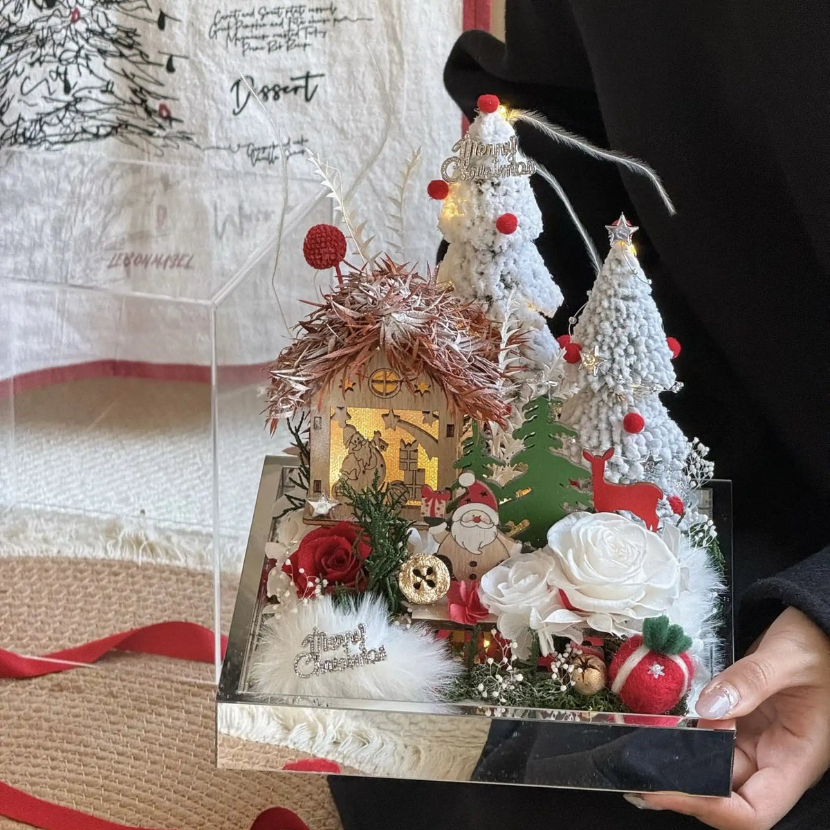 A person holds a Christmas cottage floral display with red and white preserved roses.