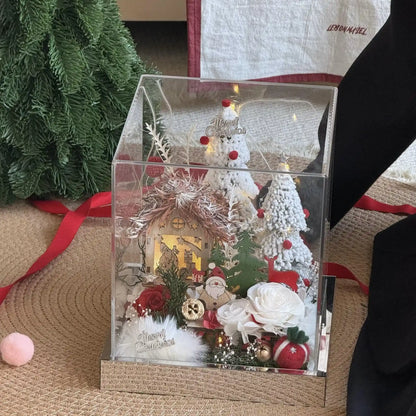 Preserved rose Christmas cottage arrangement in a clear box beside festive greenery.