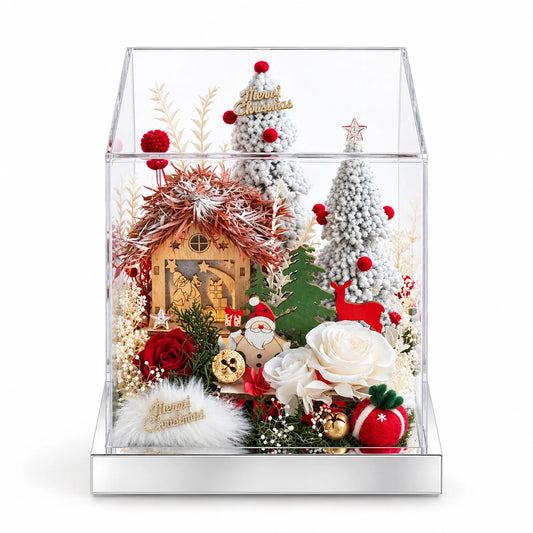 Preserved rose Christmas village with a snowy cottage and miniature trees in a clear display box on a white background.