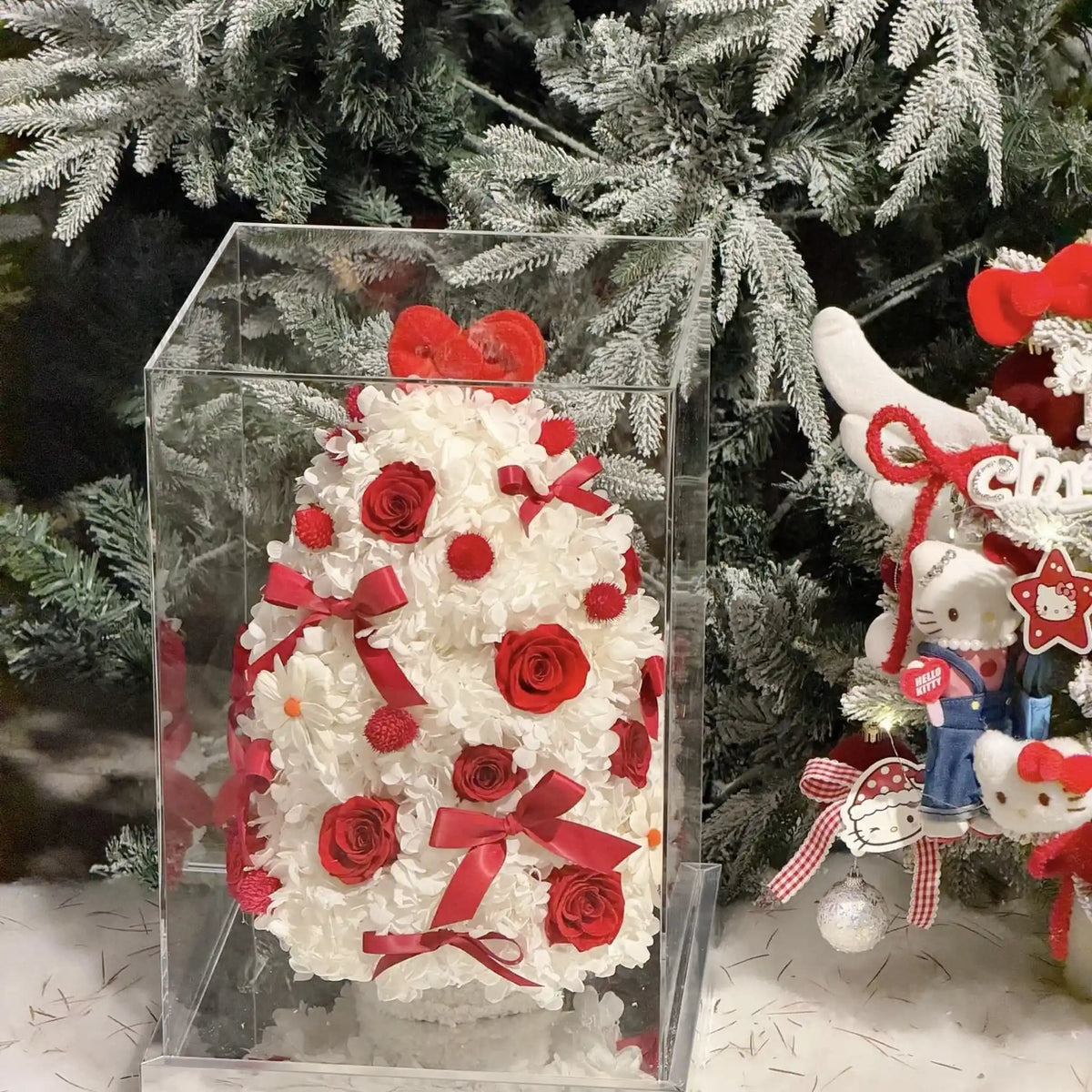 Red and white preserved flower tree in a clear box beside festive ornaments.