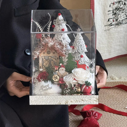 Front view of a preserved rose snow village in a clear box held by hand.