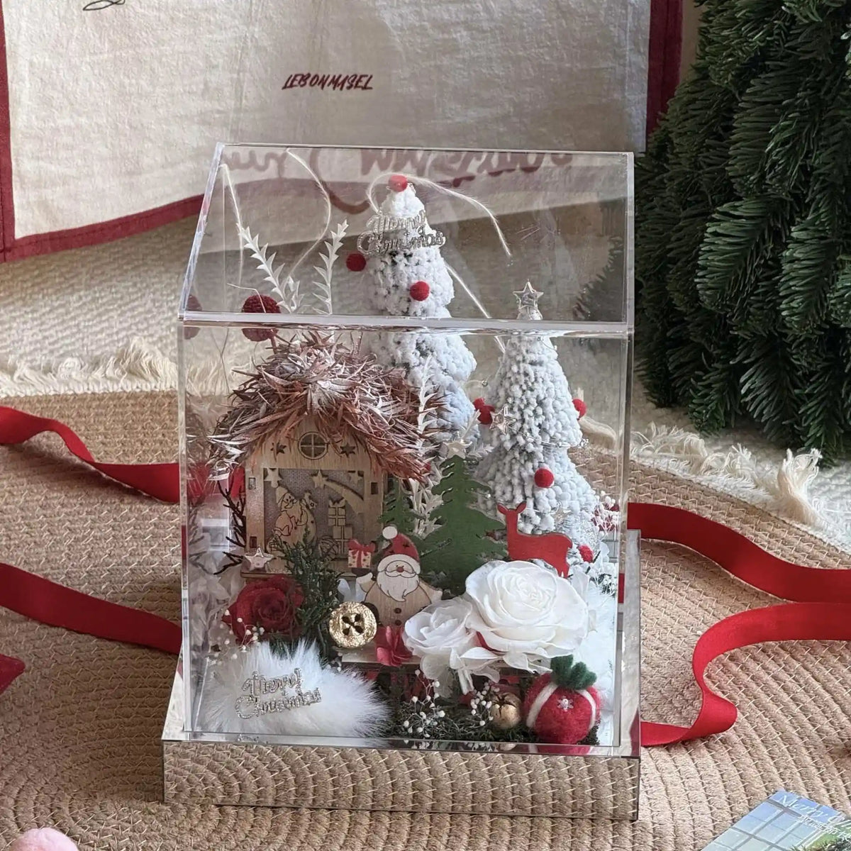 Clear boxed Christmas village with preserved roses, snowy trees and a red ribbon nearby.
