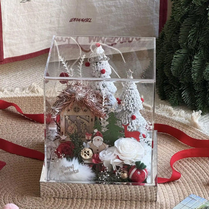Clear boxed Christmas village with preserved roses, snowy trees and a red ribbon nearby.