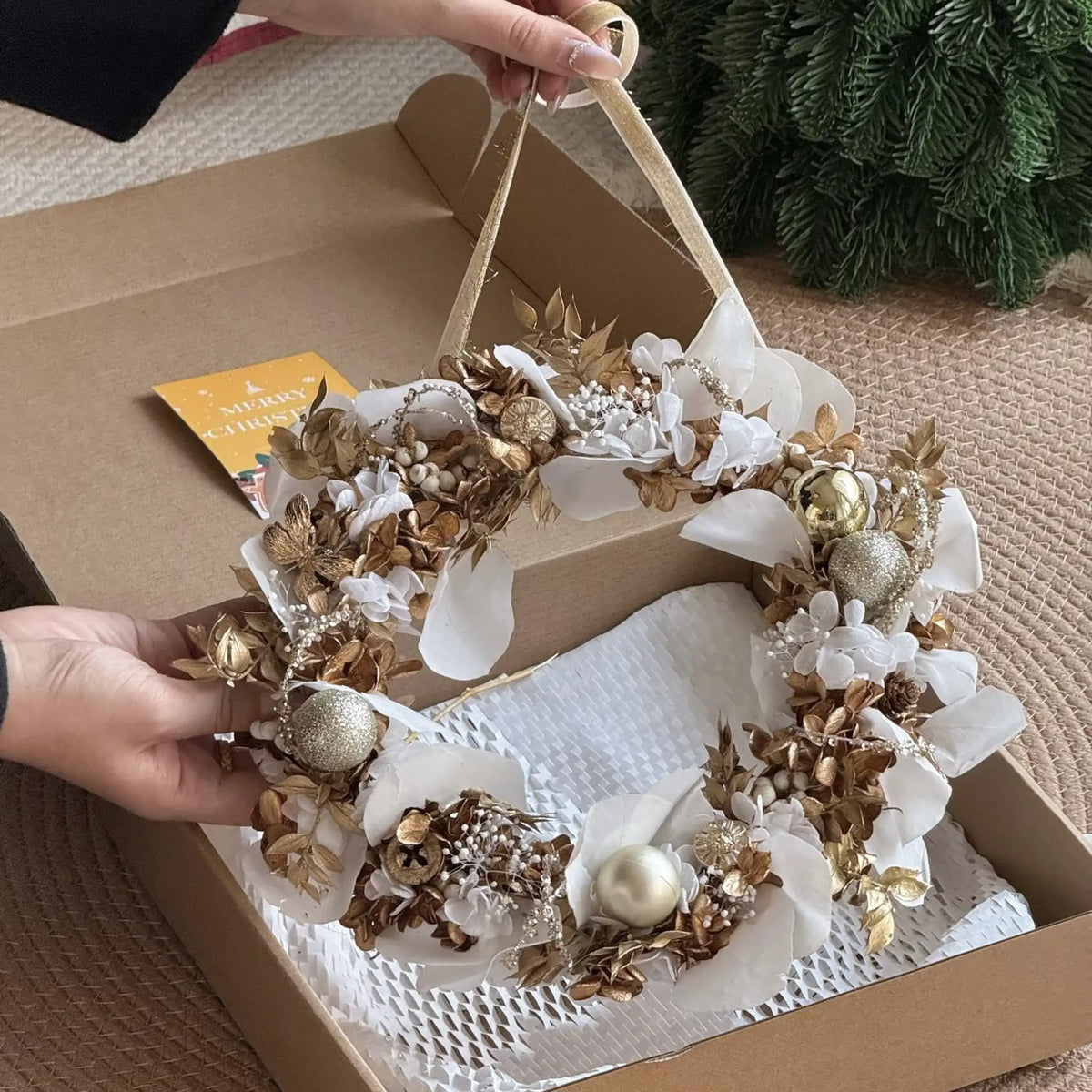 A person holds a white and gold preserved orchid and hydrangea wreath above its open gift box.