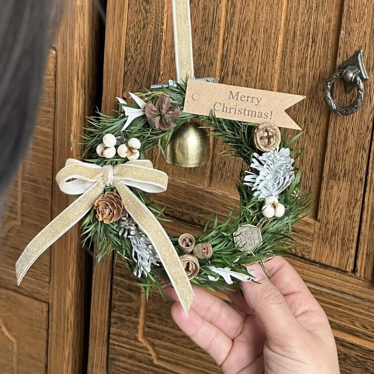 A person holds a miniature Christmas wreath with a gold bell and bow by a wooden door.