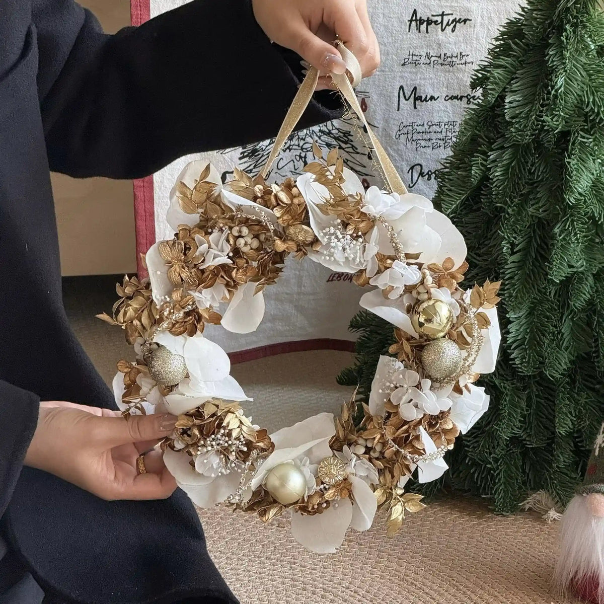 White and gold preserved flower wreath held beside an evergreen tree.