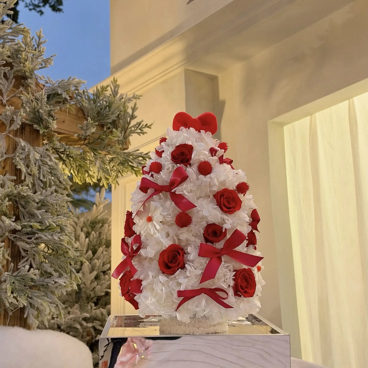 Preserved hydrangea and red rose Christmas tree displayed indoors beside festive greenery.