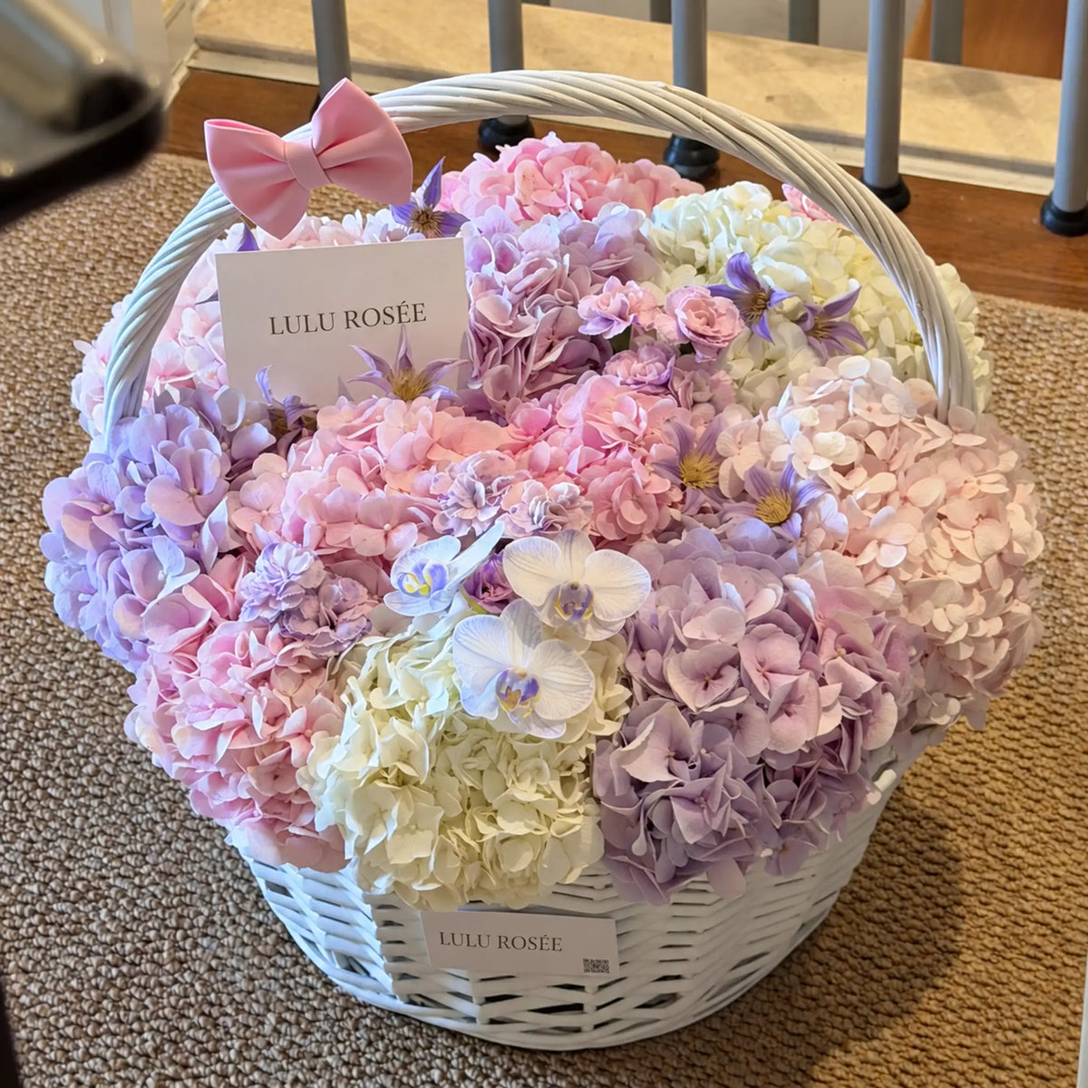 Angled view of a pastel hydrangea basket accented with white orchids and a LULU ROSÉE card