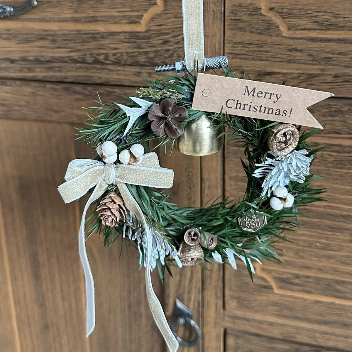 Miniature green Christmas wreath with a gold bell displayed against a wooden door.