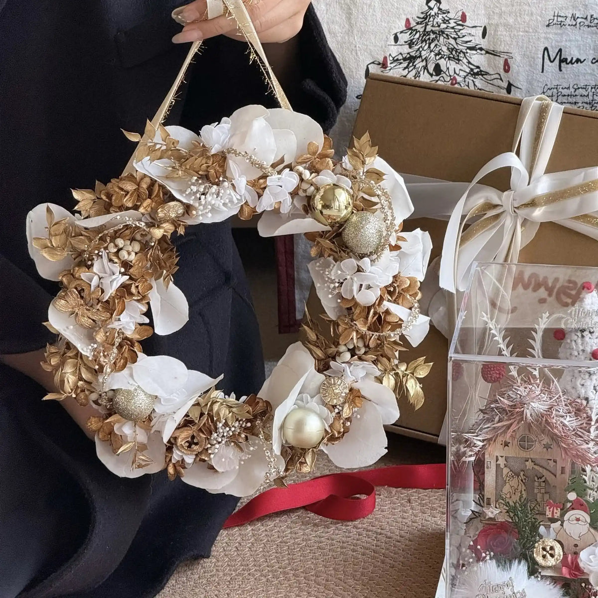 Preserved orchid and hydrangea wreath with gold ornaments displayed beside a gift box and festive scene.