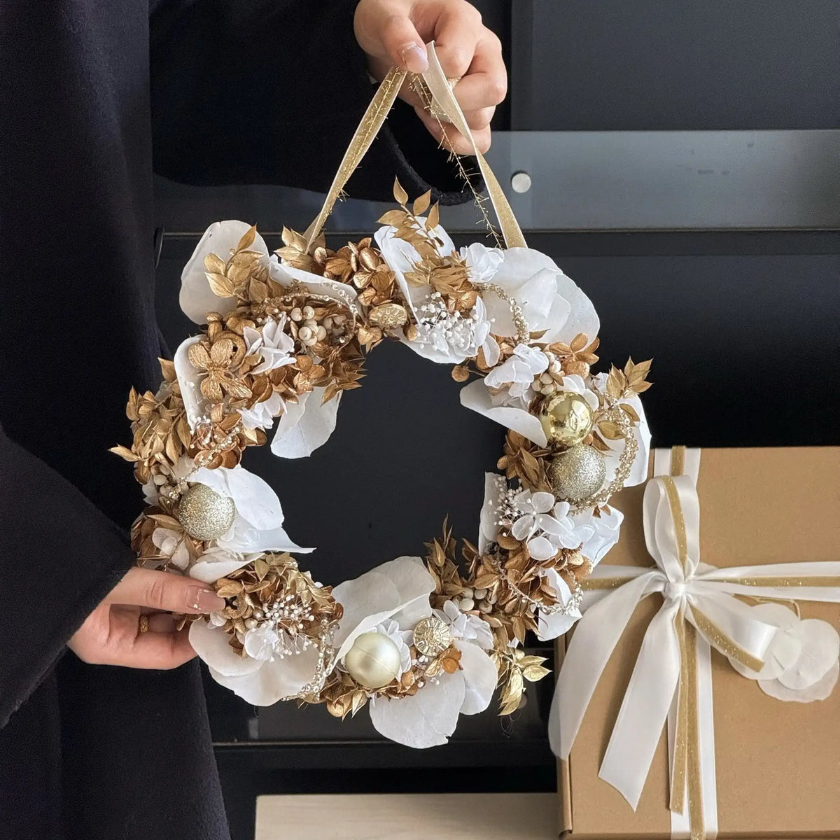 A person holds a white and gold preserved orchid and hydrangea wreath beside a ribbon-tied gift box.
