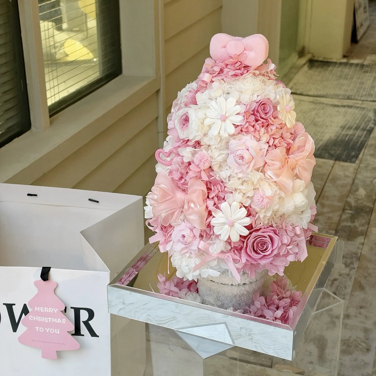 Pink preserved hydrangea and rose Christmas tree displayed outdoors on a reflective base.