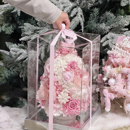 A person holding a clear gift box with a pink ribbon and preserved flower Christmas tree.